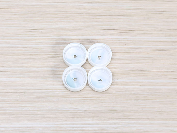 Four white plastic bottle caps are arranged in a square on a light woodgrain surface. Each cap has a small hole near the center.