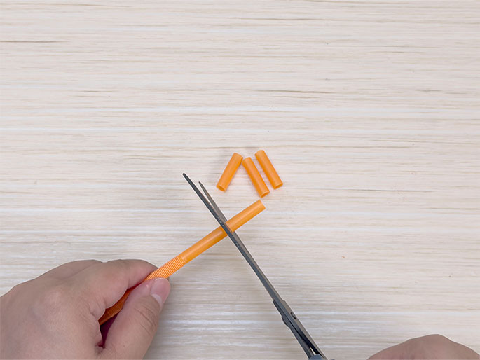 A pair of hands is using scissors to cut an orange plastic drinking straw on a light woodgrain surface. Three small pieces of orange straw already cut off lie near the top of the image.