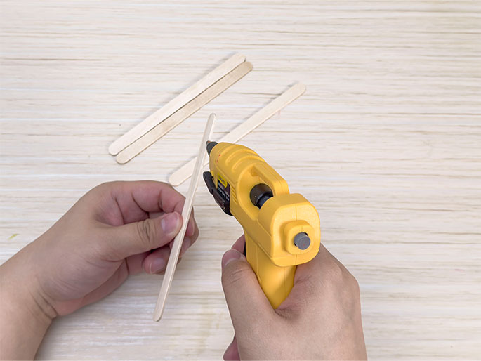 Two hands are working with craft materials on a light woodgrain surface. One hand holds a wooden craft stick, while the other hand holds a yellow hot glue gun and applies glue to the stick. Two additional craft sticks lie on the surface above. This image likely depicts the process of assembling a craft project.
