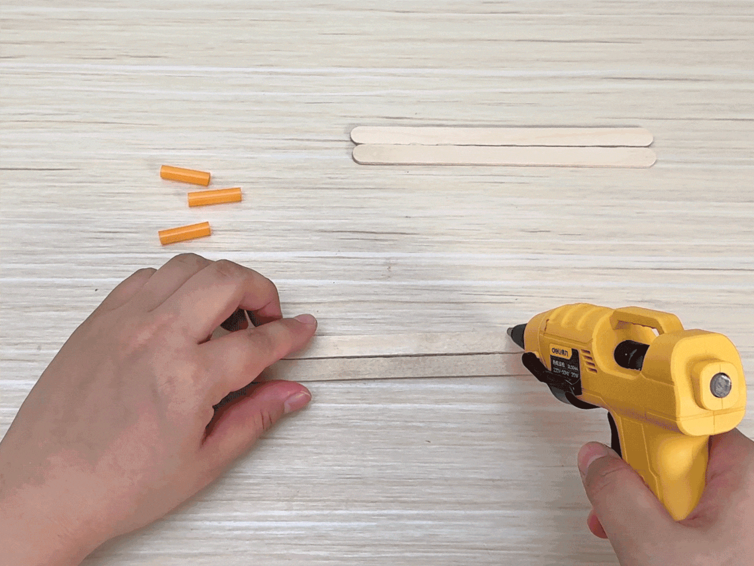 Two hands work on a craft project on a light wooden surface. Three small orange plastic pieces and two wooden craft sticks are visible in the top portion of the frame. One hand holds a wooden craft stick while the other hand uses a yellow hot glue gun to apply glue along its length. The video focuses on the process of applying the glue, presumably in preparation for attaching another piece to the stick.