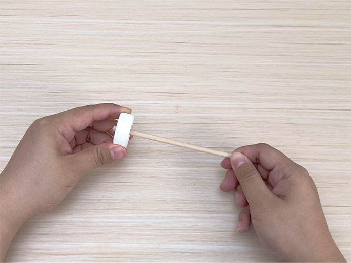 Two hands hold a small, simple construction made from a white plastic bottle cap and a thin wooden dowel on a light woodgrain surface. One hand holds the bottle cap, while the other holds the dowel, which appears to be inserted into the cap. This image likely shows a stage in a craft project.