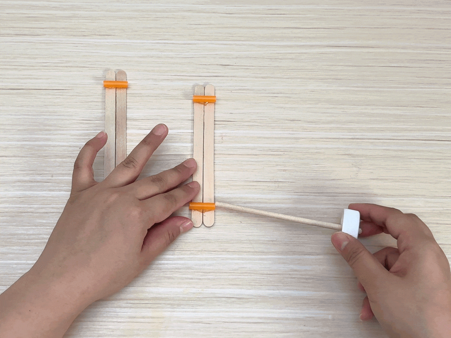 Two hands assemble a small structure on a light wooden table using popsicle sticks and small orange plastic pieces. Two pairs of popsicle sticks are laid parallel to each other, each pair bound together at the top by an orange piece. One hand positions the sticks while the other hand applies a small white plastic cap to the end of a thin wooden stick, possibly a skewer. This skewer is then inserted between the popsicle stick pairs. The video demonstrates a step in a crafting process, likely building a miniature model or toy.