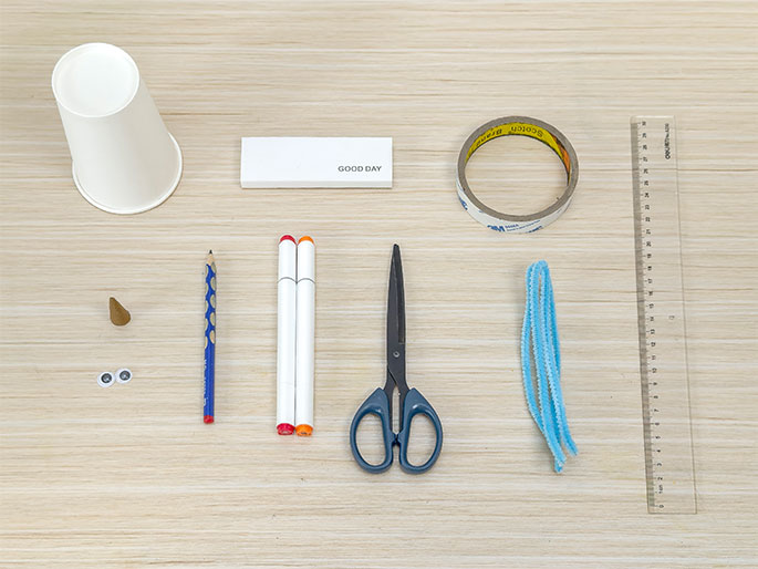 An assortment of craft supplies lies on a light wooden table, arranged for a crafting project. From left to right, the items include a white paper cup, a rectangular eraser labeled GOOD DAY, a roll of double-sided tape, a clear plastic ruler, a small brown cone shape (possibly a nose for a craft figure), two googly eyes, a blue pencil, two white markers with orange and red caps, a pair of scissors with blue handles, three light blue pipe cleaners, and a clear plastic ruler. The image displays the materials needed for a children's craft activity.