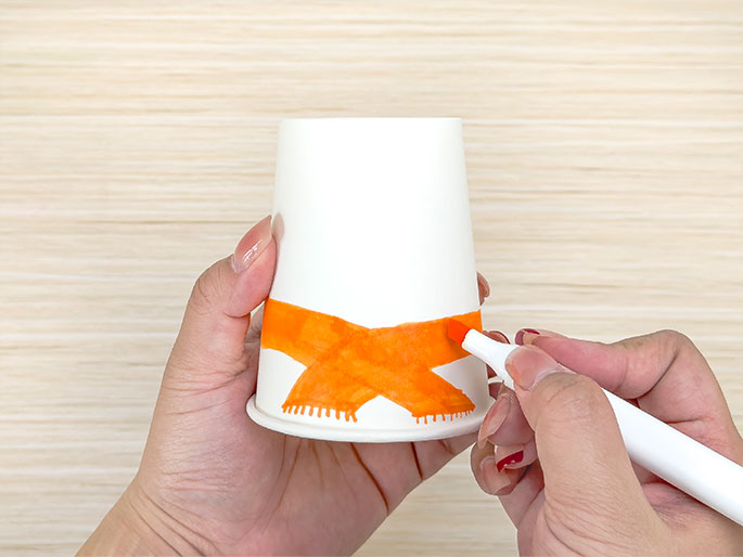 Against a light wood background, two hands are decorating a white paper cup. One hand holds the cup, which has an orange scarf design drawn around the base. Short, vertical lines extend downwards from the scarf, adding a fringe effect. The other hand holds an orange marker and is actively coloring in a section of the scarf. The image captures the process of creating a simple craft.