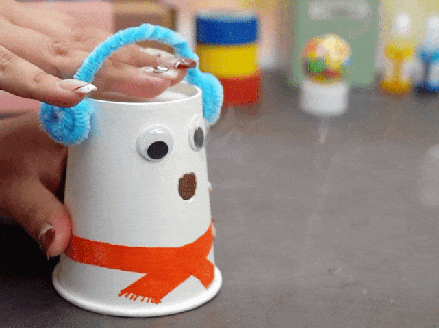 A hand interacts with a snowman craft made from a white paper cup. The snowman has googly eyes, an orange painted scarf, and blue pipe cleaner earmuffs. A small stream of smoke or vapor is being gently blown or emitted from the top of the cup, suggesting a hidden element inside. The background is blurred and includes various craft supplies.