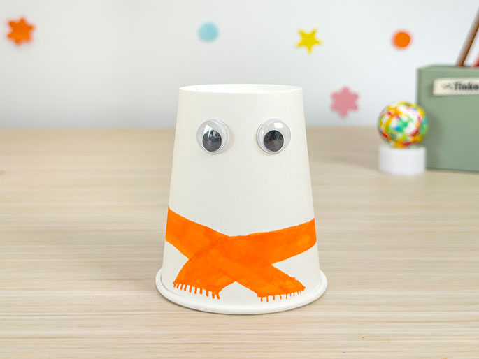 A white paper cup craft sits on a light wood table, decorated to resemble a simple snowman. Two large googly eyes are glued to the upper part of the cup. An orange scarf is drawn around the lower section, with small vertical lines indicating fringe. In the background, blurred slightly, are colorful star and circle shapes on a white wall. A small, colorful ball sits on a white pedestal, next to a light green box.