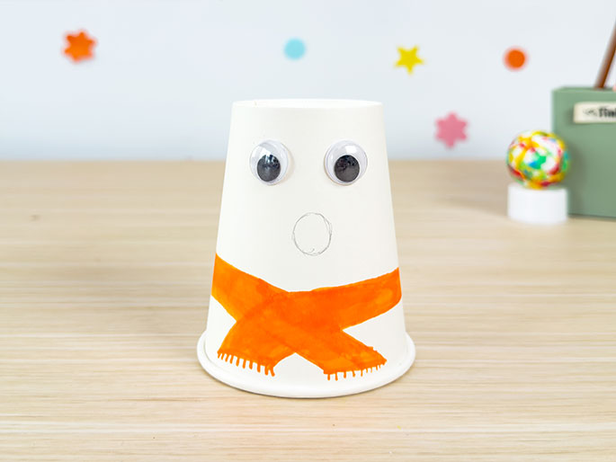 A white paper cup snowman craft rests on a light wood surface. It has two googly eyes near the top, a lightly drawn circle for a mouth, and a painted orange scarf with fringed edges around the bottom. The background features a white wall decorated with out-of-focus colorful stars and circles. A blurred colorful ball on a small white stand and a light green box are also visible in the background.