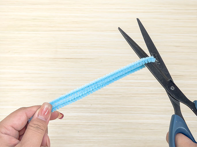 Two hands are shown against a light wooden background. One hand holds a light blue pipe cleaner, while the other hand holds a pair of scissors with blue handles, positioned to cut the pipe cleaner. The image captures the moment just before the pipe cleaner is cut, during a crafting activity.