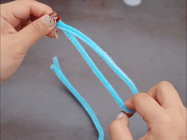 Two hands manipulate light blue pipe cleaners against a plain gray background. One hand holds two pipe cleaners together, while the other hand holds a single, slightly longer pipe cleaner. The image appears to depict a stage in a crafting process, likely preparing the pipe cleaners for assembly.