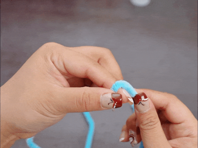 Two hands are working with a light blue pipe cleaner against a solid gray background. The fingers are folding or bending the pipe cleaner near the top. The fingernails have nail art designs.