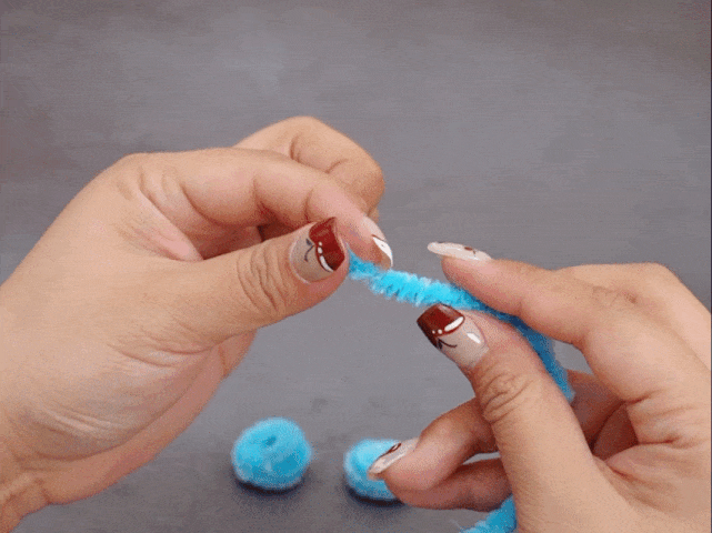 Two hands hold a light blue pipe cleaner against a gray background. The hands are pinching and twisting the pipe cleaner. Two small, fuzzy light blue pom-poms are visible in the lower left corner, suggesting they are part of the craft project. The fingernails have nail art.