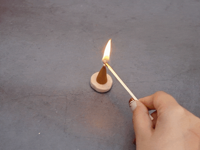 A hand holding a lit match is lighting the tip of a small, cone-shaped incense burner. The incense cone is light brown and sits on a small, round, off-white base. The background is a textured gray surface, possibly concrete or stone.