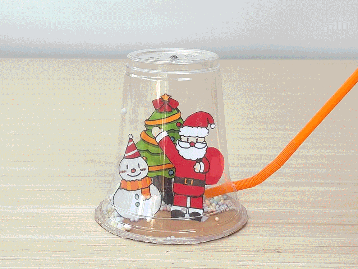 A homemade Christmas snow globe, crafted from a clear plastic cup, sits on a wooden table. Inside the cup, cartoon images of Santa Claus, a snowman, and a decorated Christmas tree are visible. Small, colorful beads, representing snow, are scattered around the figures. An orange straw is inserted into a hole near the bottom of the cup. The video showcases a festive DIY craft project, suitable for children during the holiday season.