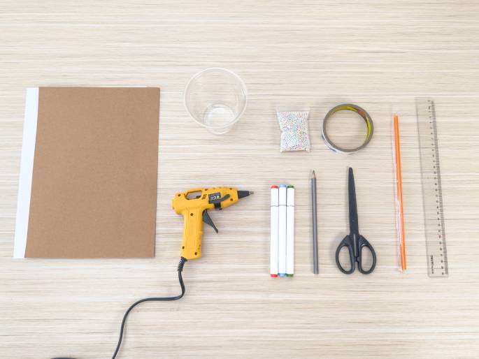 An assortment of craft supplies lies arranged on a light wood grain table. The items include a sheet of brown cardboard, a clear plastic cup, a small bag of colorful beads or sprinkles, a roll of clear tape, a yellow hot glue gun, a set of white markers, a pencil, black scissors, an orange stick or dowel, and a clear plastic ruler. This flat lay suggests the materials needed for a DIY or craft project.