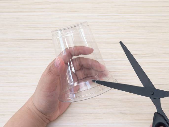A hand holds a clear plastic cup on a light wood-grain table. Another hand uses black scissors to make a small cut in the bottom rim of the cup. This suggests the start of a crafting project involving repurposing the plastic cup.