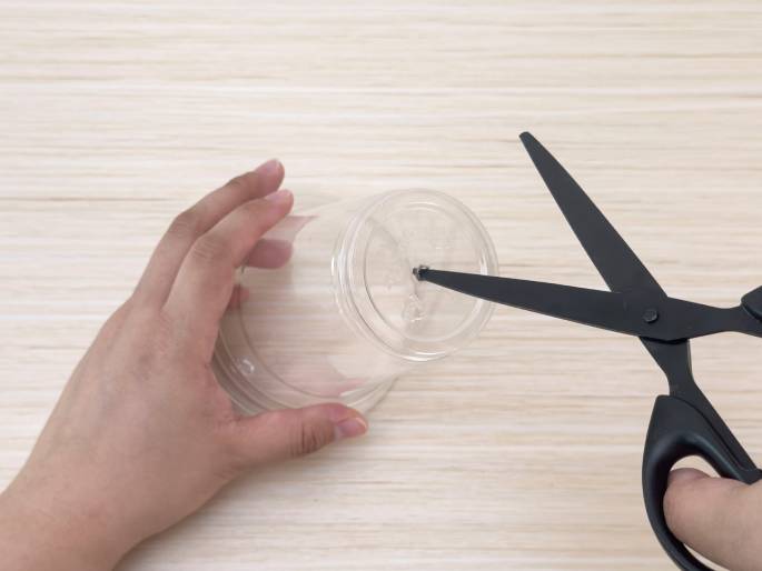 A pair of hands is working with a clear plastic cup on a light wood-grain table. One hand holds the cup upside down while the other hand uses black scissors to make a small cut in the center of the cup's bottom. This action suggests the preparation of the cup for a craft or DIY project.