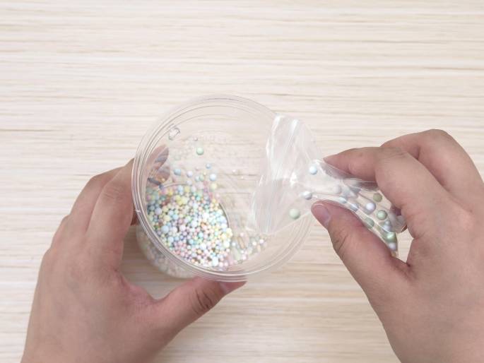Two hands are shown above a light wood-grain table. One hand holds a clear plastic container partially filled with small, pastel-colored beads. The other hand holds a clear plastic bag containing slightly larger, iridescent beads and is pouring them into the container. This suggests a crafting activity involving mixing different types of beads.