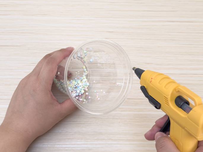 Above a light wood-grain surface, two hands manipulate craft supplies. One hand holds a clear plastic container filled with small, pastel-colored beads. The other hand operates a yellow hot glue gun, directing the nozzle towards the rim of the container, presumably to attach something to it. This scene suggests a crafting project is underway.