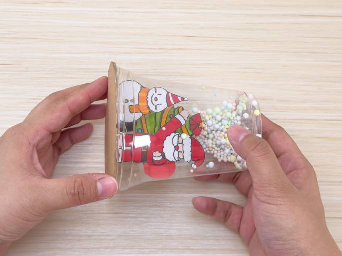 Two hands hold a clear plastic cup on its side above a light wood-grain table. Inside the cup are small, pastel-colored beads and a paper cutout of Santa Claus, a Christmas tree, and a snowman. A circular piece of brown cardboard is being attached to the open end of the cup, seemingly to seal the contents inside, creating a festive snow globe-like decoration.