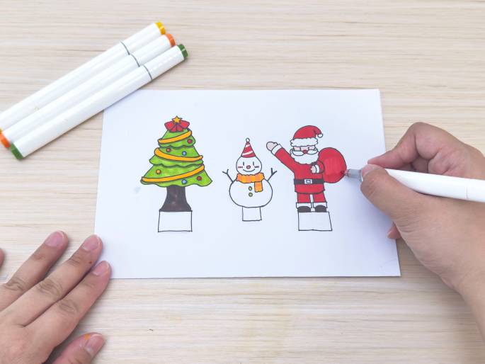 On a light wood grain table, a piece of white paper displays simple drawings of a Christmas tree, a snowman, and Santa Claus. Each drawing has a small, rectangular base. A hand holding a white marker is coloring Santa's bag red. Three white markers with orange, green, and yellow caps lie near the top left corner of the paper. This image depicts a crafting or coloring activity with a Christmas theme.