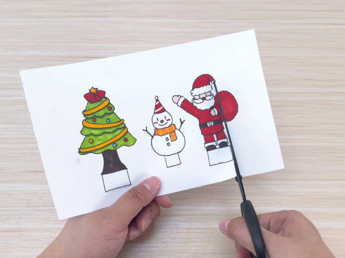 Two hands are engaged in a crafting activity on a light wood grain table. One hand holds a piece of white paper with colored drawings of a Christmas tree, a snowman, and Santa Claus. The other hand holds black scissors and is in the process of cutting out the Santa Claus figure. This image suggests creating Christmas-themed cutouts.