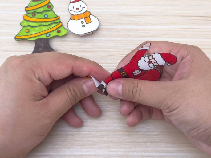 Two hands manipulate small paper cutouts of Christmas figures on a light wooden table. A completed cutout of a Christmas tree and a snowman are visible in the background. The hands are folding the rectangular base of a Santa Claus cutout. This image captures a step in a Christmas craft project, likely creating finger puppets or similar decorations.
