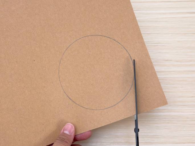 A hand holds a piece of brown cardboard with a pencil-drawn circle on it, positioned on a light wood-grain table. Black scissors are starting to cut along the line of the circle. This image indicates a crafting step where a circular shape is being cut out of the cardboard.