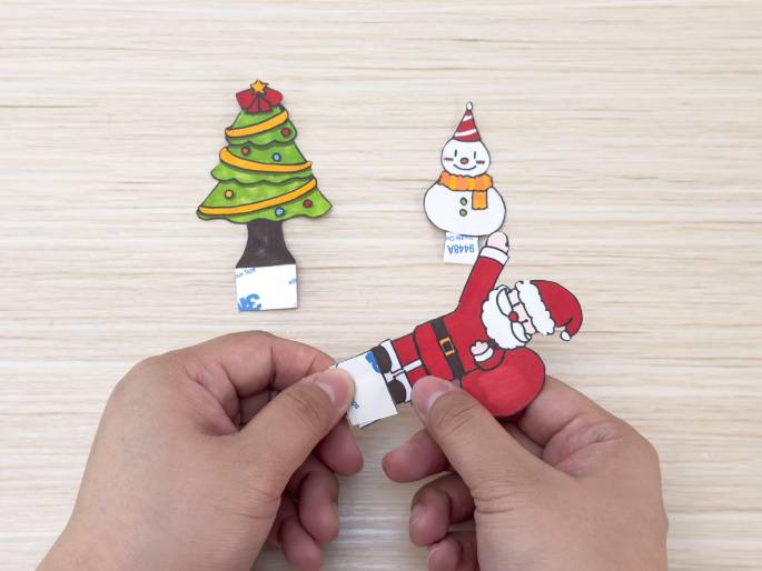 On a light wood-grain surface, two hands are preparing Christmas-themed paper cutouts. A decorated Christmas tree and a snowman are visible in the background, each with double-sided adhesive squares attached. The hands are currently peeling the backing off an adhesive square attached to a Santa Claus cutout, indicating the figures are about to be affixed to a surface as decorations.
