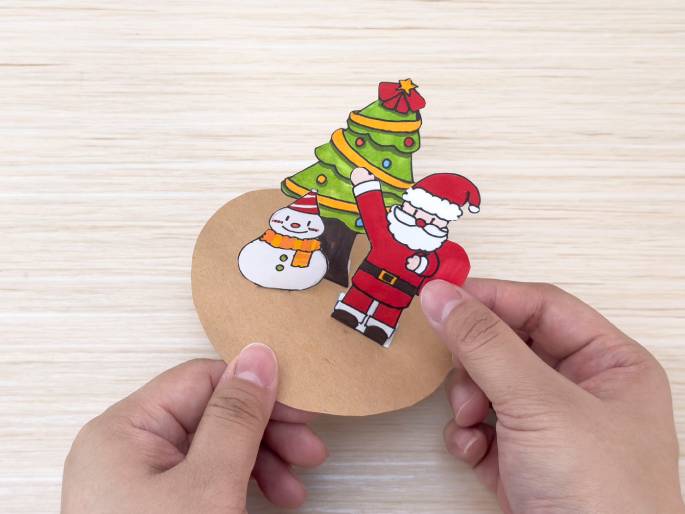 Two hands are assembling a Christmas decoration on a light wood-grain table. They are placing a cutout of Santa Claus next to a Christmas tree and a snowman. All three figures are attached to a circular piece of brown cardboard. The arrangement suggests the creation of a festive holiday ornament or card.