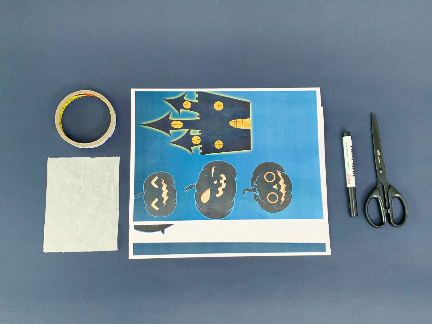 A roll of tape, a small piece of white fabric, a sheet of paper with a Halloween-themed design (castle and pumpkins), a black marker pen, and a pair of scissors are arranged on a dark blue surface.