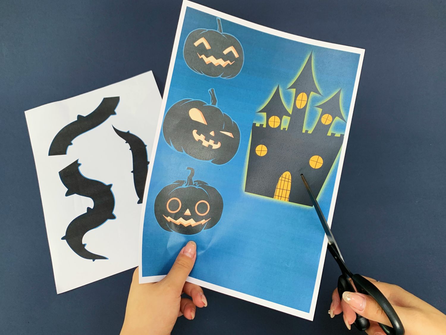 A pair of hands uses scissors to cut out a Halloween-themed design from a sheet of paper featuring a haunted house and pumpkins; another sheet shows separate spooky shapes.
