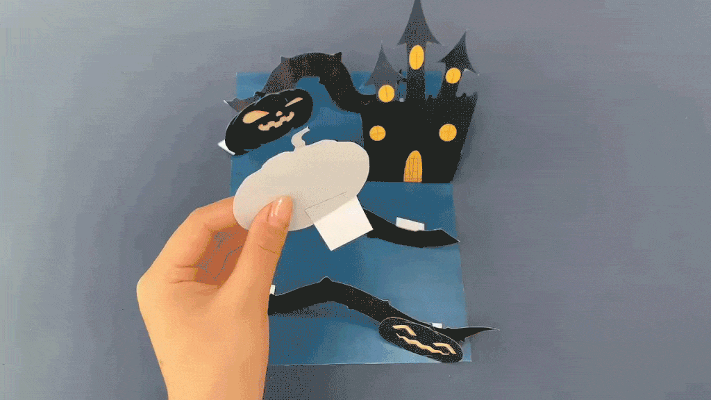 Two hands assemble a pop-up Halloween card featuring a spooky castle, pumpkins, and bats against a dark blue background.