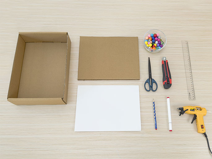 A flat lay image on a light wood background displays various craft supplies laid out, seemingly in preparation for a project. The materials include an open cardboard box, a flat piece of cardboard, a sheet of white paper, a small bowl of colorful beads, scissors, a utility knife, a ruler, a pencil, a white marker, and a hot glue gun. The arrangement suggests the items will be used together for a crafting or DIY activity.