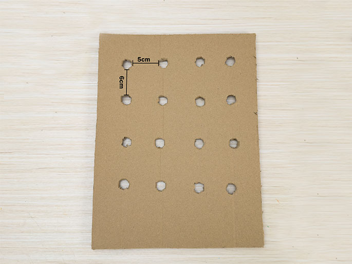 A rectangular piece of light brown cardboard lies on a light wood-grained surface. The cardboard has a grid of evenly spaced holes punched in it. Lines and text indicate the spacing between the holes: 6cm vertically and 5cm horizontally. This appears to be a prepared piece for a craft or game.