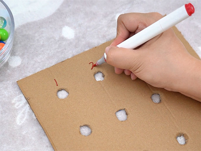 A hand holding a white marker with a red cap is writing the number 2 next to a hole in a piece of light brown cardboard. The cardboard has a grid of holes and rests on a textured white surface. A small bowl of colorful beads is partially visible in the top left corner. The number 1 is already written next to another hole, suggesting the holes are being numbered sequentially. This appears to be a step in a craft or game-making process.