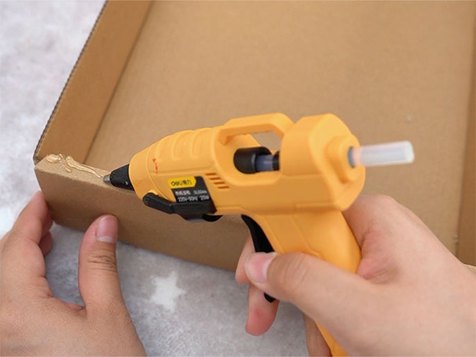 Hands are using a bright orange hot glue gun to apply glue to the inside edge of a cardboard box. The glue is a light tan color and is being dispensed onto the cardboard. The box and hands rest on a white textured surface. This image appears to depict a step in a crafting or DIY project.