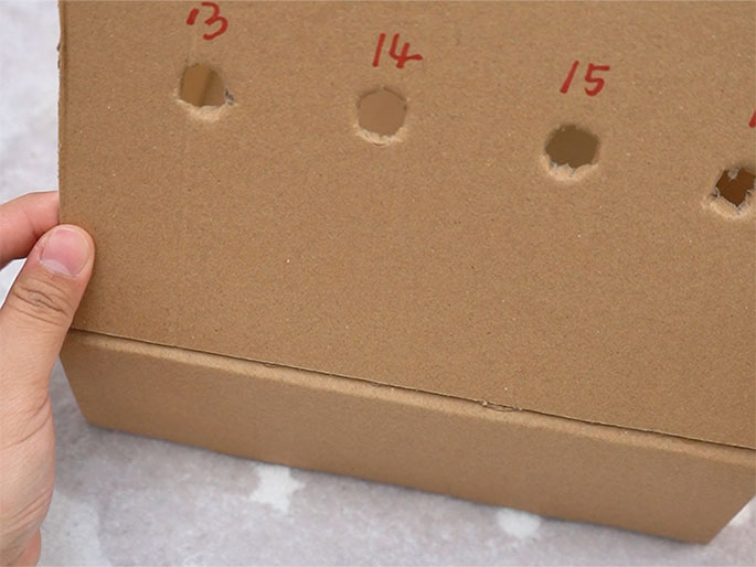 A piece of cardboard with punched holes and numbers written in red ink next to them (13, 14, 15, and a partially visible 16) is being attached to a cardboard box. A hand is visible on the left side, holding the cardboard piece in place against the box. The box and hand are positioned on a textured white surface. This image seems to depict a stage in a DIY or crafting project.