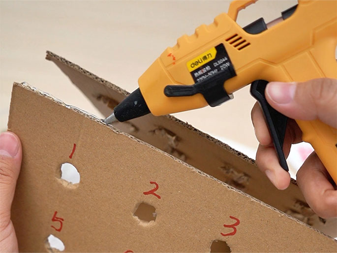 A hand uses a bright orange hot glue gun to apply glue to the edge of a piece of cardboard. The cardboard has holes punched in it and numbers written in red (1, 2, 3, and 5 are visible). Another piece of cardboard is positioned below the first, suggesting the glue is being used to attach them together. This appears to be a step in constructing a game or craft project.