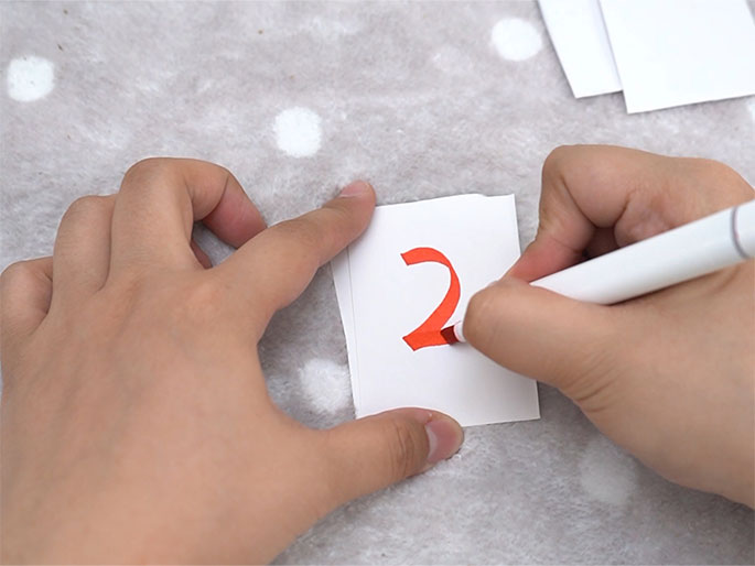 Two hands are shown on a textured white surface with small white polka dots. One hand holds a small white card while the other hand uses a white marker with a red cap to write the number 2 in red ink on the card. A small stack of similar blank white cards is visible in the upper right corner. This image appears to be of someone preparing numbered cards for a game or craft activity.