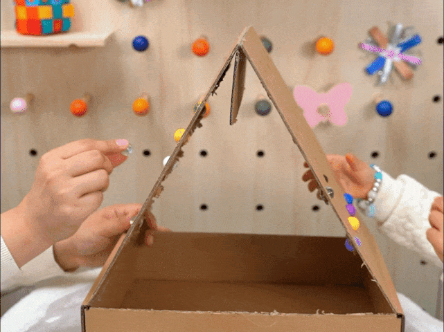 Two hands interact with a cardboard structure shaped like a triangular prism, reminiscent of a simple house. Small, colorful balls are placed into holes along the angled sides of the structure. The hands adjust the position of the structure, causing the balls to roll down the inside and collect at the bottom. The background features a pegboard decorated with colorful pegs and a paper butterfly. This video likely depicts a simple, homemade toy or game involving dropping balls into a cardboard structure.