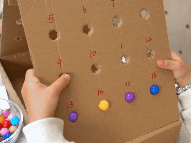 Small hands place colorful gumballs into numbered holes on a piece of cardboard. The cardboard appears to be part of a homemade game or activity. Numbers are written next to each hole, presumably corresponding to the quantity or order of gumballs to be placed. A bowl filled with more colorful gumballs sits nearby. The video focuses on the close-up interaction of the hands placing the gumballs into the designated holes.