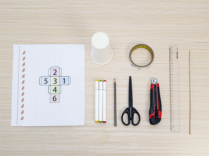 A flat lay arrangement of craft supplies on a light wood grain surface. The supplies include a printed sheet of paper with a cube net (a flattened cube design with numbers 1-6) and a decorative dotted border, a white paper cup, a roll of clear tape, a ruler, a thin wooden dowel, a pair of scissors, a utility knife, a pencil, and two white markers with green and red caps. These items suggest preparation for a craft or DIY project involving paper and potentially other materials.