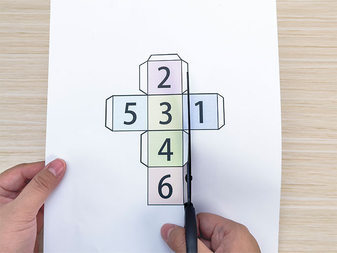 A pair of hands is cutting out a printed paper cube net. The net is laid out flat on a light wooden surface and has the numbers 1 through 6 printed on its faces, each in a different pastel color. The scissors are cutting along one of the lines of the template. This image shows the process of creating a paper cube, likely for a game or educational activity.