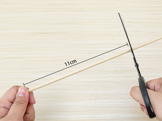 Hands are using scissors to cut an 11cm long wooden dowel or stick. The background is a light woodgrain surface. The image focuses on the action of cutting the dowel, likely for a craft or construction project.
