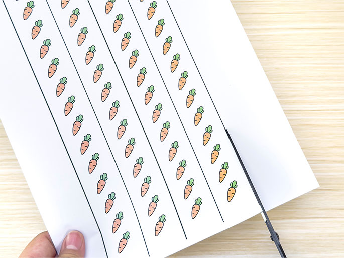 A hand holds a piece of white paper with rows of printed carrot illustrations against a light wooden background. Scissors are positioned to cut the paper, likely to separate the carrot rows. This image suggests a step in a crafting activity, perhaps preparing elements for a game or decoration.
