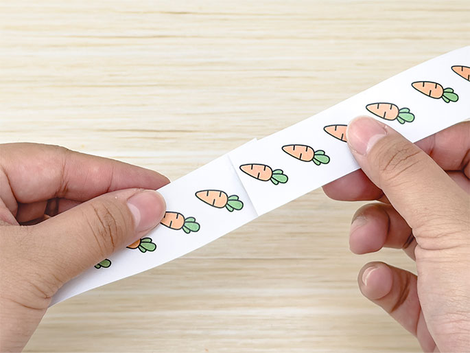Two hands are folding a strip of white paper printed with a row of cartoon carrots. The paper is being folded between each carrot image, creating a zig-zag or accordion-style fold. The background is a light wooden surface. This action likely represents a step in a craft project.