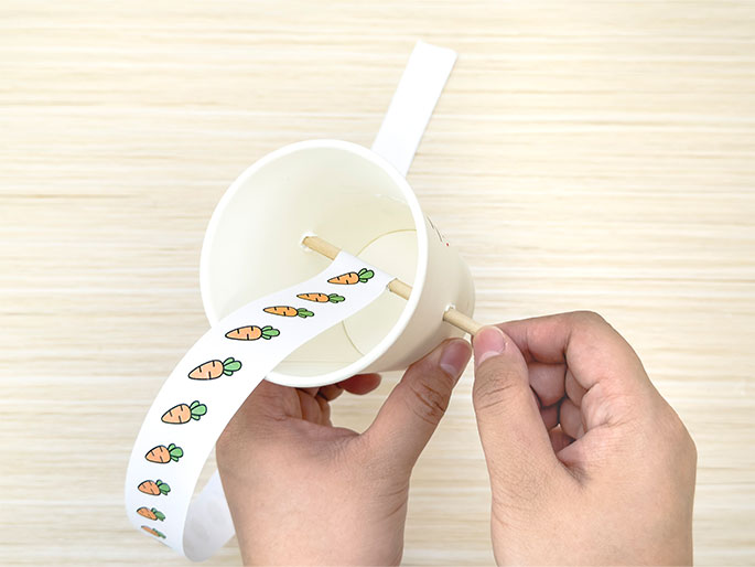 Two hands are assembling a craft project involving a white paper cup, a thin wooden dowel, and a strip of paper printed with carrots. The dowel is inserted through holes on opposite sides of the cup, and the carrot-printed paper strip is wrapped around the dowel inside the cup. The image is taken against a light woodgrain background and shows a stage in the construction of what might be a toy or decoration.