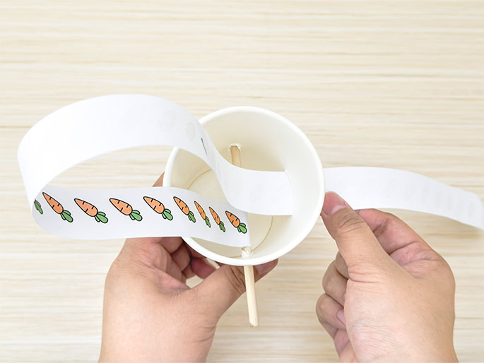 Against a light woodgrain background, two hands are shown threading a strip of paper decorated with carrots through a white paper cup. A small wooden dowel or stick is already inserted through holes in the cup, and the paper strip is being looped over and around it. This image captures a step in creating a simple craft, possibly a toy or decorative item.