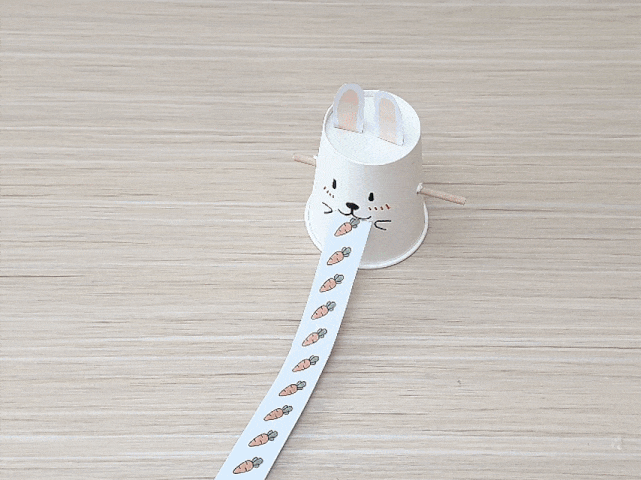 A paper cup, decorated with a cute bunny face and ears, sits on a light wooden surface. A long strip of paper, printed with small carrot images, is being pulled out from the bunny’s mouth. Small wooden sticks protrude from the sides of the cup, likely serving as arms. This video likely demonstrates a simple craft or toy, possibly designed for children, showcasing a bunny “eating” a paper strip of carrots.