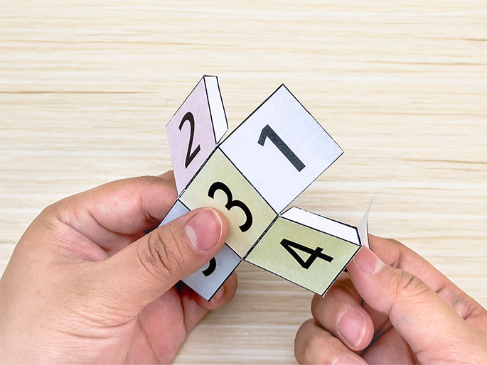 Two hands manipulate a partially assembled paper cube net on a light wood background. The net is printed with the numbers 1 to 6 in different pastel-colored squares. The hands are folding the net, and some of the joining tabs are visible. The image captures the process of constructing a paper cube.